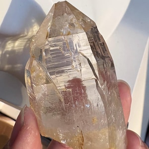 Puede incluir: Un espécimen de cristal de cuarzo claro y puntiagudo con una estructura hexagonal y fracturas internas visibles. El cristal presenta una base translúcida de color marrón dorado pálido y facetas transparentes afiladas que captan la luz, mostrando estrías geométricas naturales.