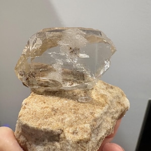Puede incluir: Un grupo de cristales de cuarzo transparentes y claros con inclusiones internas y facetas visibles, fijado sobre una base de matriz mineral rugosa y texturizada de color beige claro. El cristal presenta un brillo v&iacute;treo y formas geom&eacute;tricas irregulares.