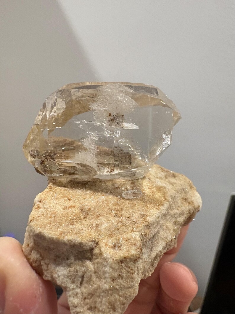 Puede incluir: Un grupo de cristales de cuarzo transparentes y claros con inclusiones internas y facetas visibles, fijado sobre una base de matriz mineral rugosa y texturizada de color beige claro. El cristal presenta un brillo v&iacute;treo y formas geom&eacute;tricas irregulares.