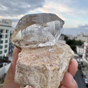 Puede incluir: Un cristal de cuarzo transparente y facetado unido a una matriz mineral rugosa de color beige. El cristal presenta fracturas internas y estr&iacute;as visibles, con una apariencia transparente y v&iacute;trea que contrasta con la base terrosa y texturizada.