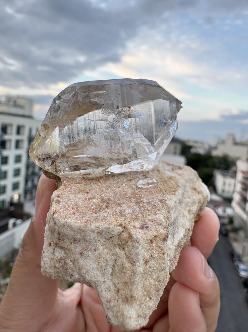 Puede incluir: Un cristal de cuarzo transparente y facetado unido a una matriz mineral rugosa de color beige. El cristal presenta fracturas internas y estr&iacute;as visibles, con una apariencia transparente y v&iacute;trea que contrasta con la base terrosa y texturizada.