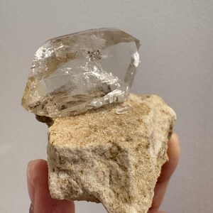 Puede incluir: Un cristal de cuarzo transparente y transl&uacute;cido con facetas angulares afiladas unido a una matriz mineral rugosa y texturizada de color tostado claro. El cristal muestra inclusiones internas y un brillo v&iacute;treo que contrasta con la roca base mate y terrosa.