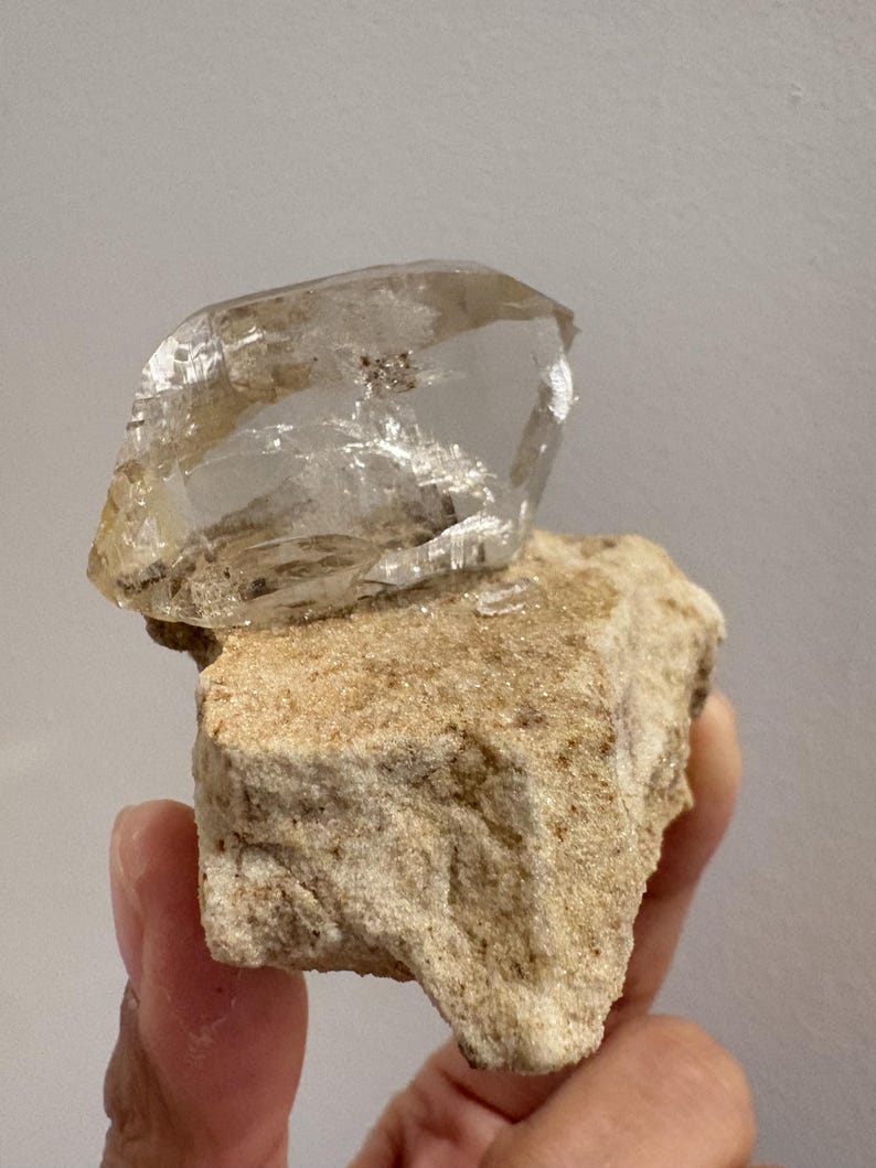 Puede incluir: Un cristal de cuarzo transparente y transl&uacute;cido con facetas angulares afiladas unido a una matriz mineral rugosa y texturizada de color tostado claro. El cristal muestra inclusiones internas y un brillo v&iacute;treo que contrasta con la roca base mate y terrosa.