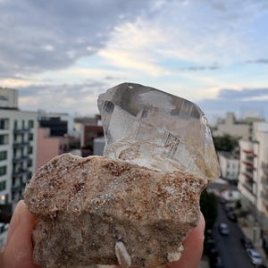 Puede incluir: Una punta de cristal de cuarzo transparente unida a una base de matriz mineral rugosa, texturizada y de color marr&oacute;n claro. El cristal presenta facetas geom&eacute;tricas distintas e inclusiones internas, contrastando con la superficie terrosa y granular de la roca.