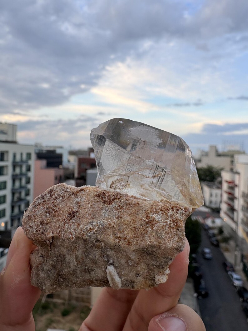 Puede incluir: Una punta de cristal de cuarzo transparente unida a una base de matriz mineral rugosa, texturizada y de color marr&oacute;n claro. El cristal presenta facetas geom&eacute;tricas distintas e inclusiones internas, contrastando con la superficie terrosa y granular de la roca.