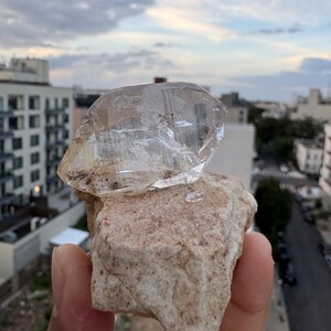 Puede incluir: Una punta de cristal de cuarzo transparente y facetada unida a una base de matriz mineral rugosa de color beige. El cristal presenta estr&iacute;as naturales e inclusiones internas, mientras que la base tiene una superficie texturizada e irregular con peque&ntilde;as motas marrones.