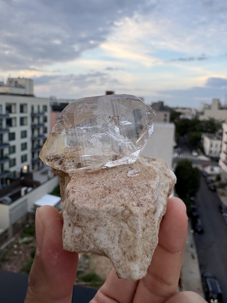 Puede incluir: Una punta de cristal de cuarzo transparente y facetada unida a una base de matriz mineral rugosa de color beige. El cristal presenta estr&iacute;as naturales e inclusiones internas, mientras que la base tiene una superficie texturizada e irregular con peque&ntilde;as motas marrones.
