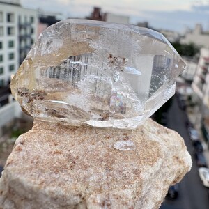 Puede incluir: Un esp&eacute;cimen de cristal de cuarzo transparente y facetado con inclusiones internas y una peque&ntilde;a refracci&oacute;n de arco&iacute;ris, que descansa sobre una base de matriz mineral rugosa y texturizada de color tostado claro con detalles granulares visibles.