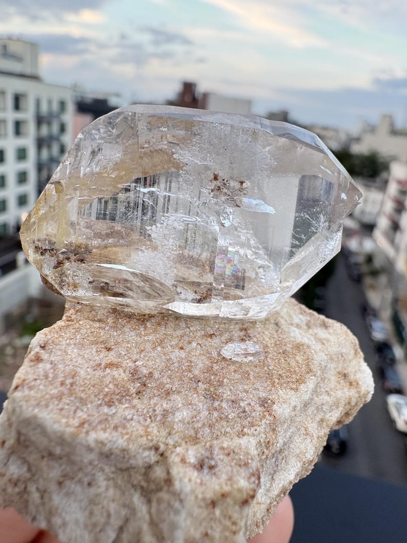 Puede incluir: Un esp&eacute;cimen de cristal de cuarzo transparente y facetado con inclusiones internas y una peque&ntilde;a refracci&oacute;n de arco&iacute;ris, que descansa sobre una base de matriz mineral rugosa y texturizada de color tostado claro con detalles granulares visibles.