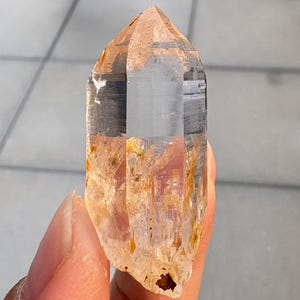 Puede incluir: Una punta de cristal de cuarzo hexagonal transparente con inclusiones minerales de color naranja dorado en su interior, concentradas cerca de la base y la parte central, con un ápice natural afilado y caras verticales translúcidas.