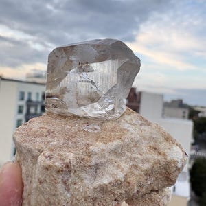 Puede incluir: Un esp&eacute;cimen de cristal de cuarzo transparente y facetado con inclusiones internas visibles, fijado sobre una base de matriz mineral rugosa y texturizada de color tostado claro. El cristal presenta bordes geom&eacute;tricos afilados y una superficie v&iacute;trea.