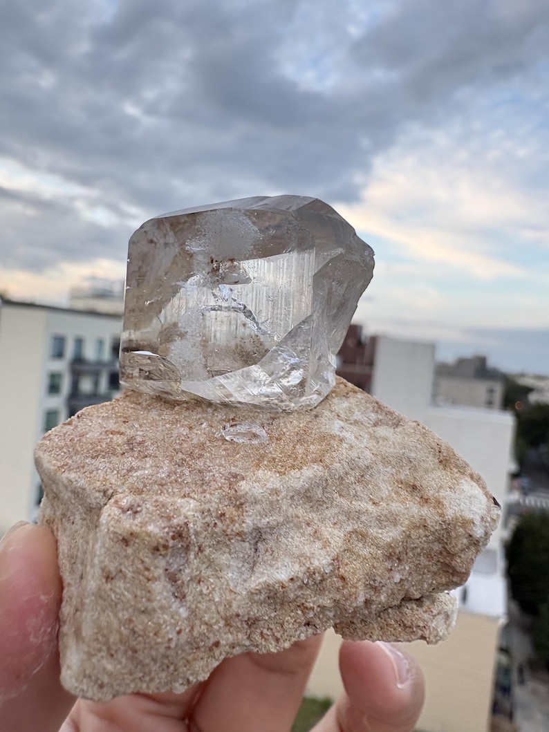 Puede incluir: Un esp&eacute;cimen de cristal de cuarzo transparente y facetado con inclusiones internas visibles, fijado sobre una base de matriz mineral rugosa y texturizada de color tostado claro. El cristal presenta bordes geom&eacute;tricos afilados y una superficie v&iacute;trea.