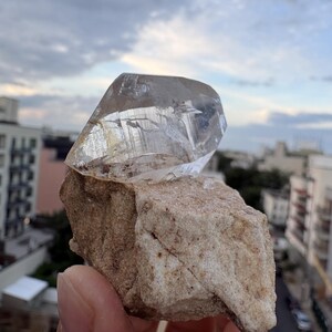Puede incluir: Una punta de cristal de cuarzo transparente y facetada unida a una base de matriz mineral de color beige rugoso. El cristal presenta estr&iacute;as internas visibles y una textura v&iacute;trea, mientras que la base tiene una superficie gruesa e irregular.