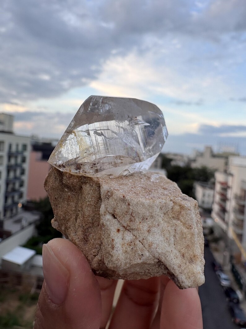 Puede incluir: Una punta de cristal de cuarzo transparente y facetada unida a una base de matriz mineral de color beige rugoso. El cristal presenta estr&iacute;as internas visibles y una textura v&iacute;trea, mientras que la base tiene una superficie gruesa e irregular.