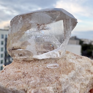 Puede incluir: Un esp&eacute;cimen de cristal de cuarzo grande, claro y multifac&eacute;tico con fracturas e inclusiones internas visibles, que descansa sobre una matriz de piedra de color beige, rugosa y texturizada, con peque&ntilde;os dep&oacute;sitos minerales de color marr&oacute;n rojizo.