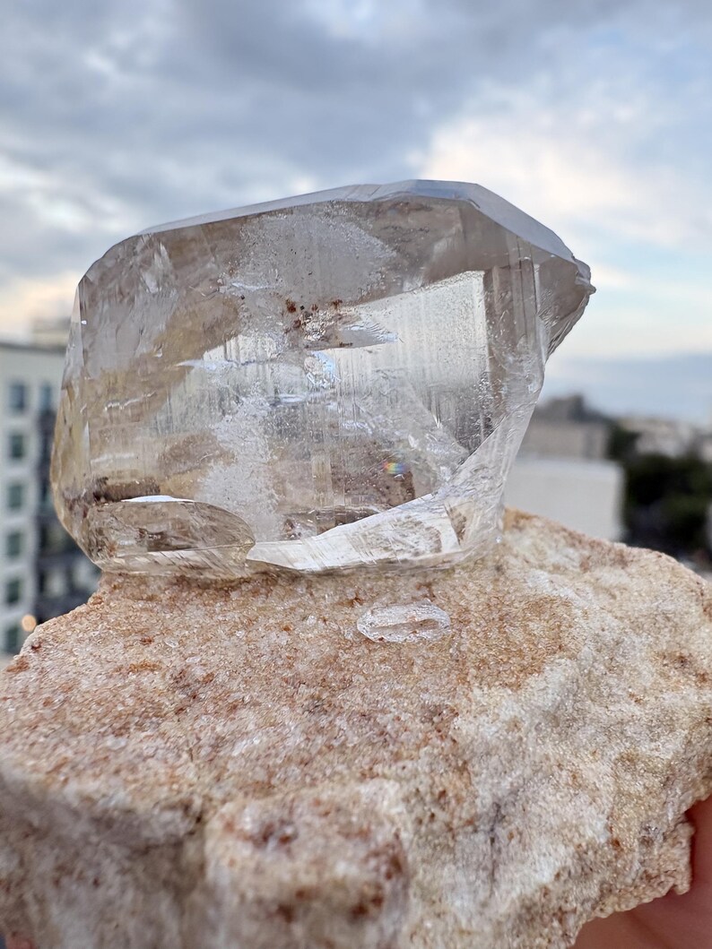 Puede incluir: Un esp&eacute;cimen de cristal de cuarzo grande, claro y multifac&eacute;tico con fracturas e inclusiones internas visibles, que descansa sobre una matriz de piedra de color beige, rugosa y texturizada, con peque&ntilde;os dep&oacute;sitos minerales de color marr&oacute;n rojizo.