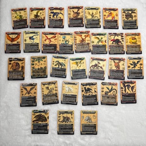 May include: A collection of twenty-eight rectangular trading cards featuring various dragon illustrations, each with detailed text blocks, stats, and unique creature names printed in black and gold borders, arranged in a grid on a white textured surface.
