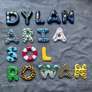May include: A collection of hand-painted letters forming the names DYLAN, ARIA, SOL, and ROWAN. Each letter features unique designs including space themes, floral patterns, animal prints, a life preserver, and a landscape with pink flamingos.