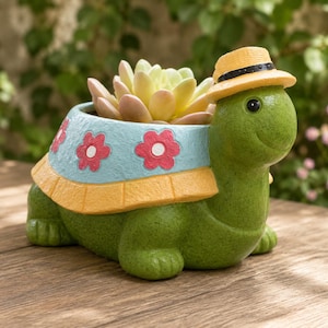 Peut inclure: Un pot de fleurs en forme de tortue verte avec une carapace bleu clair ornée de fleurs roses et d'une bordure jaune. La tortue porte un petit chapeau jaune avec un ruban noir. Une plante succulente claire est plantée dans la carapace.