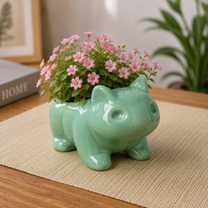 Peut inclure: Un pot de fleurs en céramique vert clair en forme de créature à quatre pattes avec des oreilles pointues et un dos creux. Le pot est rempli d'un groupe dense de petites fleurs roses éclatantes avec un feuillage vert, créant un ensemble coloré.