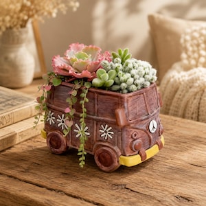 Peut inclure: Un pot de fleurs en céramique marron en forme de van vintage avec un pare-chocs jaune, des motifs de fleurs blanches et un emblème de signe de paix. Le pot contient diverses succulentes vertes et roses, dont une grande echeveria frisée.