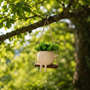 Peut inclure: Un pot de fleurs en céramique blanc cassé avec un visage souriant, des bras et des jambes, rempli de plantes vertes. Le pot est suspendu à une branche d'arbre sur une balançoire en bois, suspendue par une corde. L'arrière-plan est une vue floue du feuillage vert.