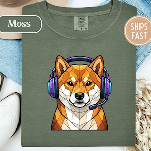May include: A moss green t-shirt featuring a geometric, low-poly style illustration of a Shiba Inu dog wearing purple and blue gaming headphones with a microphone. The shirt has a Comfort Colors brand label at the inner neckline.