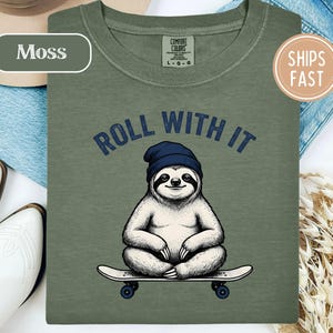 May include: A moss green crew neck t-shirt featuring a graphic of a sloth wearing a navy blue beanie and sitting on a skateboard. The text "ROLL WITH IT" is printed in dark blue above the sloth. The shirt has a Comfort Colors brand label.