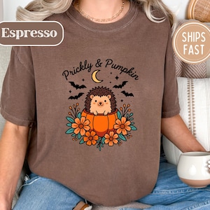 May include: A brown t-shirt featuring a graphic of a hedgehog sitting inside an orange pumpkin. The design includes orange flowers, black bats, a yellow crescent moon, and the text "Prickly & Pumpkin" printed in black above the hedgehog illustration.