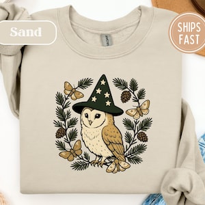 May include: A sand-colored crewneck sweatshirt featuring a graphic print of a barn owl wearing a dark green pointed hat with yellow stars. The owl is surrounded by a circular wreath of pine branches, pinecones, and four beige moths.