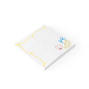 May include: A square stack of white paper sheets featuring a yellow decorative border line. The design includes a small blue bow, a pink seashell, and a yellow citrus slice illustration printed on the top sheet of the paper stack.