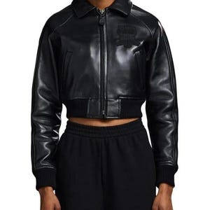 May include: A black cropped leather jacket featuring a classic collar, a full-length front zipper closure, and ribbed knit cuffs and hem. The jacket includes a tonal embroidered patch on the upper left chest and long sleeves with visible seam detailing.