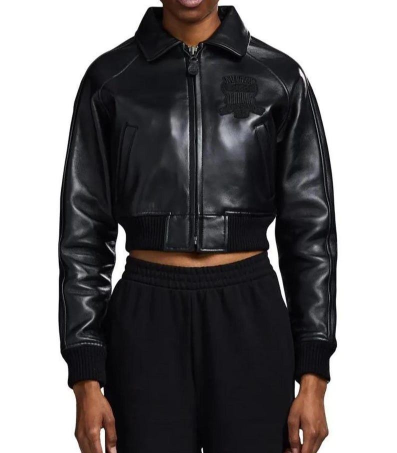 May include: A black cropped leather jacket featuring a classic collar, a full-length front zipper closure, and ribbed knit cuffs and hem. The jacket includes a tonal embroidered patch on the upper left chest and long sleeves with visible seam detailing.