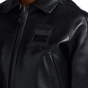 May include: A black leather jacket featuring a full-length front zipper with a branded pull tab, a classic collar, and long sleeves. An embroidered patch on the chest displays the text AVIREX ESTABLISHED 1975 with a shield and star graphic.