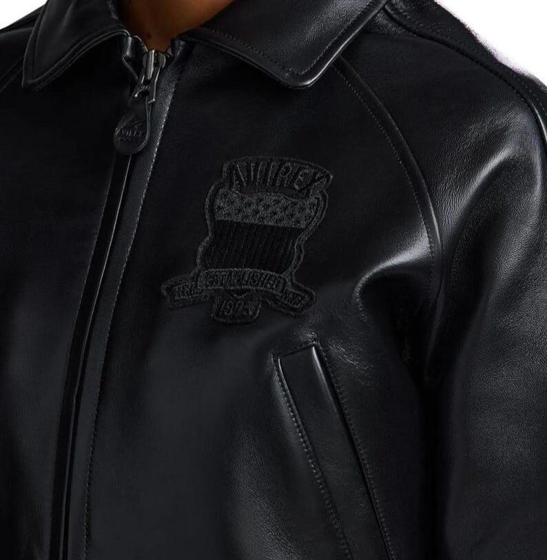 May include: A black leather jacket featuring a full-length front zipper with a branded pull tab, a classic collar, and long sleeves. An embroidered patch on the chest displays the text AVIREX ESTABLISHED 1975 with a shield and star graphic.