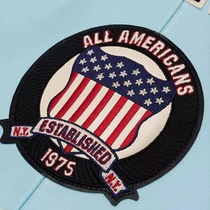 Pu&ograve; includere: Una toppa ricamata circolare su sfondo azzurro raffigurante uno scudo con strisce rosse e bianche e un campo blu con stelle bianche. Il testo ALL AMERICANS, ESTABLISHED 1975 e N.Y. &egrave; ricamato in rosso e bianco su un bordo nero.