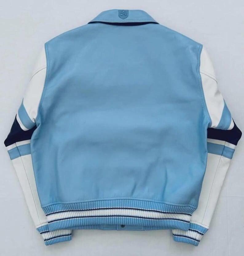 Op de afbeelding: Een lichtblauw varsity-jack met witte leren mouwen, een geribbelde kraag en gestreepte boorden in lichtblauw, wit en marineblauw. Het achterpand is effen lichtblauw met een klein reli&euml;flogo in het midden van de kraag.