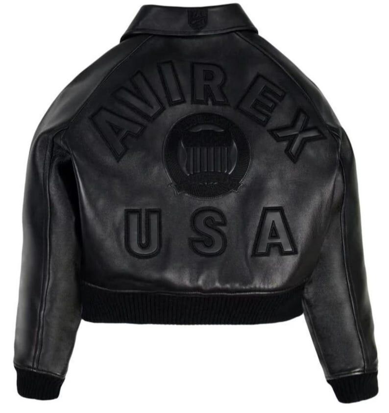 May include: A black leather jacket featuring a collar and ribbed cuffs and hem. The back displays the embossed text AVIREX USA in a curved arrangement above a circular emblem with vertical stripes, all in a matching black tone on the leather.