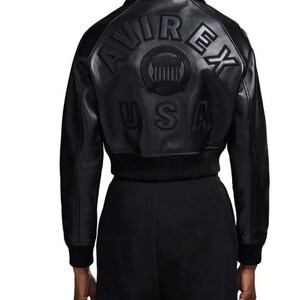 May include: A black leather bomber jacket featuring a ribbed collar, cuffs, and hem. The back displays embossed lettering that reads AVIREX USA, accompanied by a circular emblem with a shield design centered between the text and the USA label.