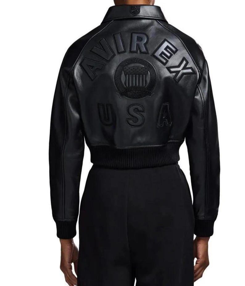 May include: A black leather bomber jacket featuring a ribbed collar, cuffs, and hem. The back displays embossed lettering that reads AVIREX USA, accompanied by a circular emblem with a shield design centered between the text and the USA label.