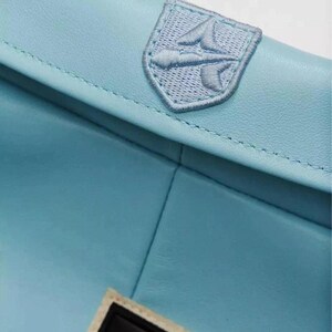 Pu&ograve; includere: Un accessorio in pelle azzurro con una superficie liscia e cuciture visibili lungo i bordi. Una toppa ricamata a forma di scudo, realizzata con filo azzurro coordinato, &egrave; centrata sulla patta e mostra un emblema stilizzato a forma di freccia.