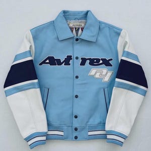 Op de afbeelding: Een lichtblauw varsity jack met witte en marineblauwe accenten. De voorzijde is voorzien van de tekst Avirex in donkerblauwe letters, een witte afbeelding van het getal 41, een puntige kraag, een knoopsluiting en geribbelde boorden.