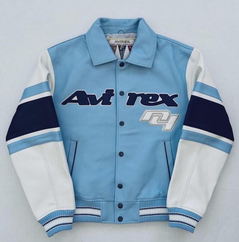 Op de afbeelding: Een lichtblauw varsity jack met witte en marineblauwe accenten. De voorzijde is voorzien van de tekst Avirex in donkerblauwe letters, een witte afbeelding van het getal 41, een puntige kraag, een knoopsluiting en geribbelde boorden.