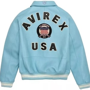 Pu&ograve; includere: Una giacca varsity azzurra con la scritta nera AVIREX arcuata sulla parte superiore della schiena. Sotto, una toppa circolare mostra il testo ALL AMERICANS ESTABLISHED 1975 con una bandiera americana. La parola USA appare in grandi lettere nere.