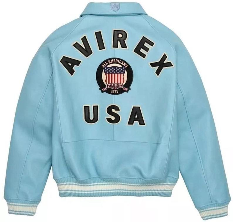 Pu&ograve; includere: Una giacca varsity azzurra con la scritta nera AVIREX arcuata sulla parte superiore della schiena. Sotto, una toppa circolare mostra il testo ALL AMERICANS ESTABLISHED 1975 con una bandiera americana. La parola USA appare in grandi lettere nere.