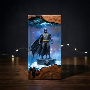 Puede incluir: Una l&aacute;mpara rectangular de resina y madera con una figura de Batman sobre una base de g&aacute;rgola. La secci&oacute;n central de resina transparente est&aacute; iluminada con una luz azul, creando un efecto de nebulosa detr&aacute;s de la figura.