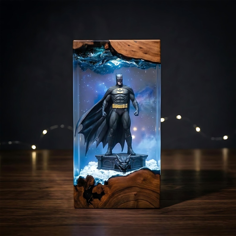Puede incluir: Una l&aacute;mpara rectangular de resina y madera con una figura de Batman sobre una base de g&aacute;rgola. La secci&oacute;n central de resina transparente est&aacute; iluminada con una luz azul, creando un efecto de nebulosa detr&aacute;s de la figura.