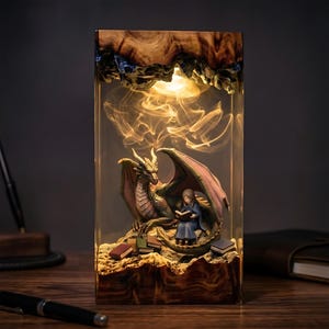 May include: A rectangular resin lamp featuring a natural wood base and top. Inside the clear resin, a sculpted dragon with spread wings sits beside a small figure reading a book. Several miniature books are scattered on the rocky base.