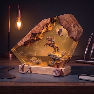 Puede incluir: Una lámpara decorativa con marco de madera pulida que encierra un bloque de resina ámbar translúcida. Dentro, una escena en miniatura muestra a un jinete sobre un caballo encabritado, un águila volando y flora del desierto. Una herradura y una pequeña bolsa de cuero descansan en la base.