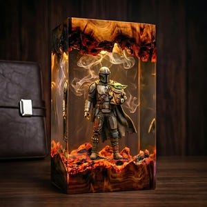 May include: A rectangular resin lamp featuring a Mandalorian figure holding a small green creature inside a clear block. The base and top are made of polished wood with natural grain, and the interior contains swirling white smoke effects.